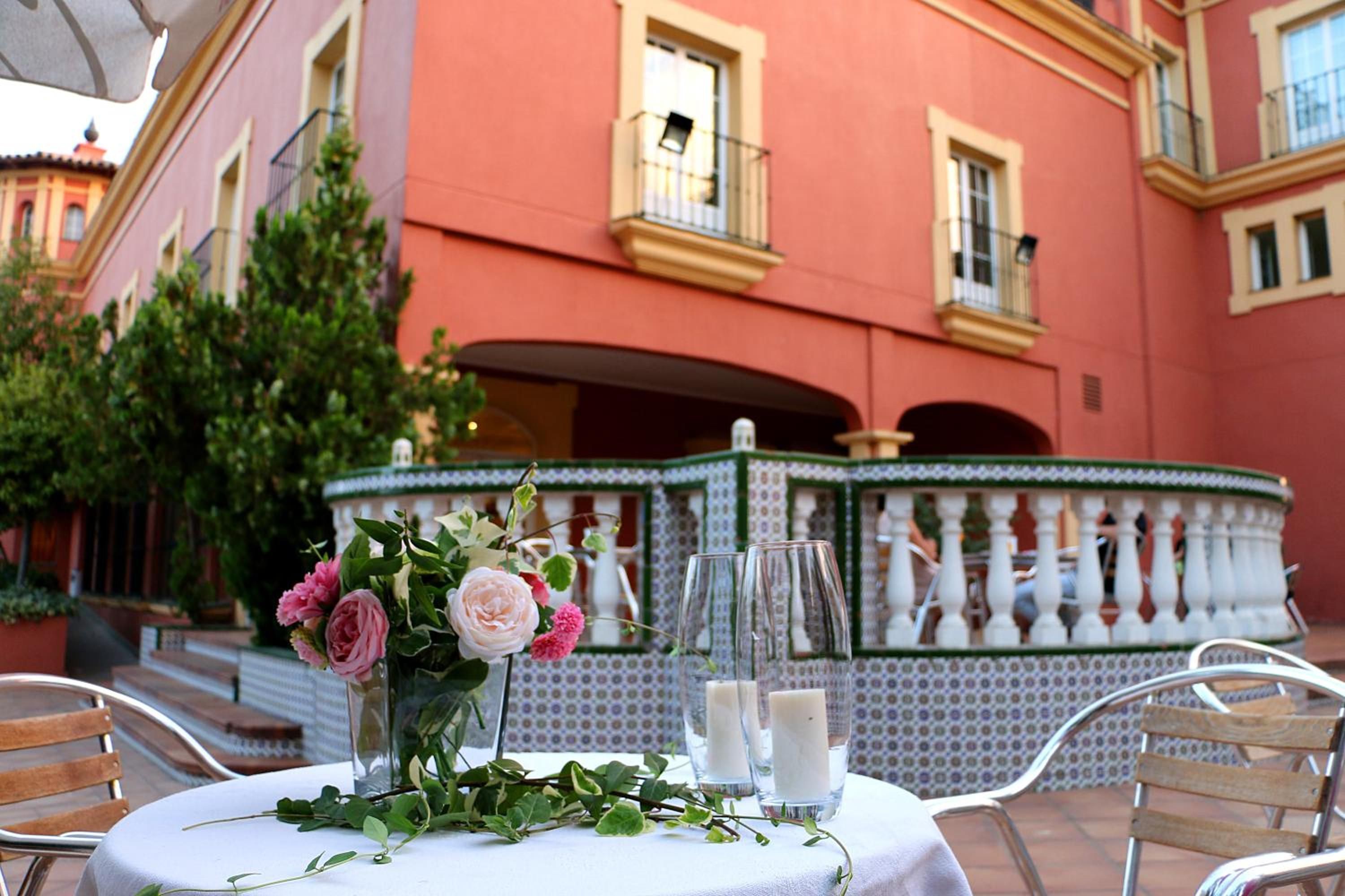 Un área de comedor al aire libre con una mesa dispuesta para dos, un jarrón decorativo con flores y una fuente al fondo.