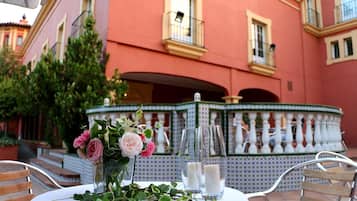 Un área de comedor al aire libre con una mesa dispuesta para dos, un jarrón decorativo con flores y una fuente al fondo.