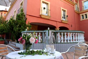 Un área de comedor al aire libre con una mesa dispuesta para dos, un jarrón decorativo con flores y una fuente al fondo.
