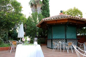 An outdoor patio with a covered seating area, a table set with a vase of flowers, and a tall tree in the background. - Hotel Cristina (Fregenal de la Sierra)