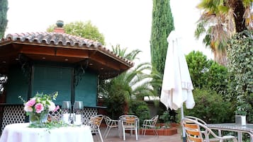 Un patio al aire libre con un área de estar cubierta, una mesa con un centro de mesa floral y una palmera alta al fondo.