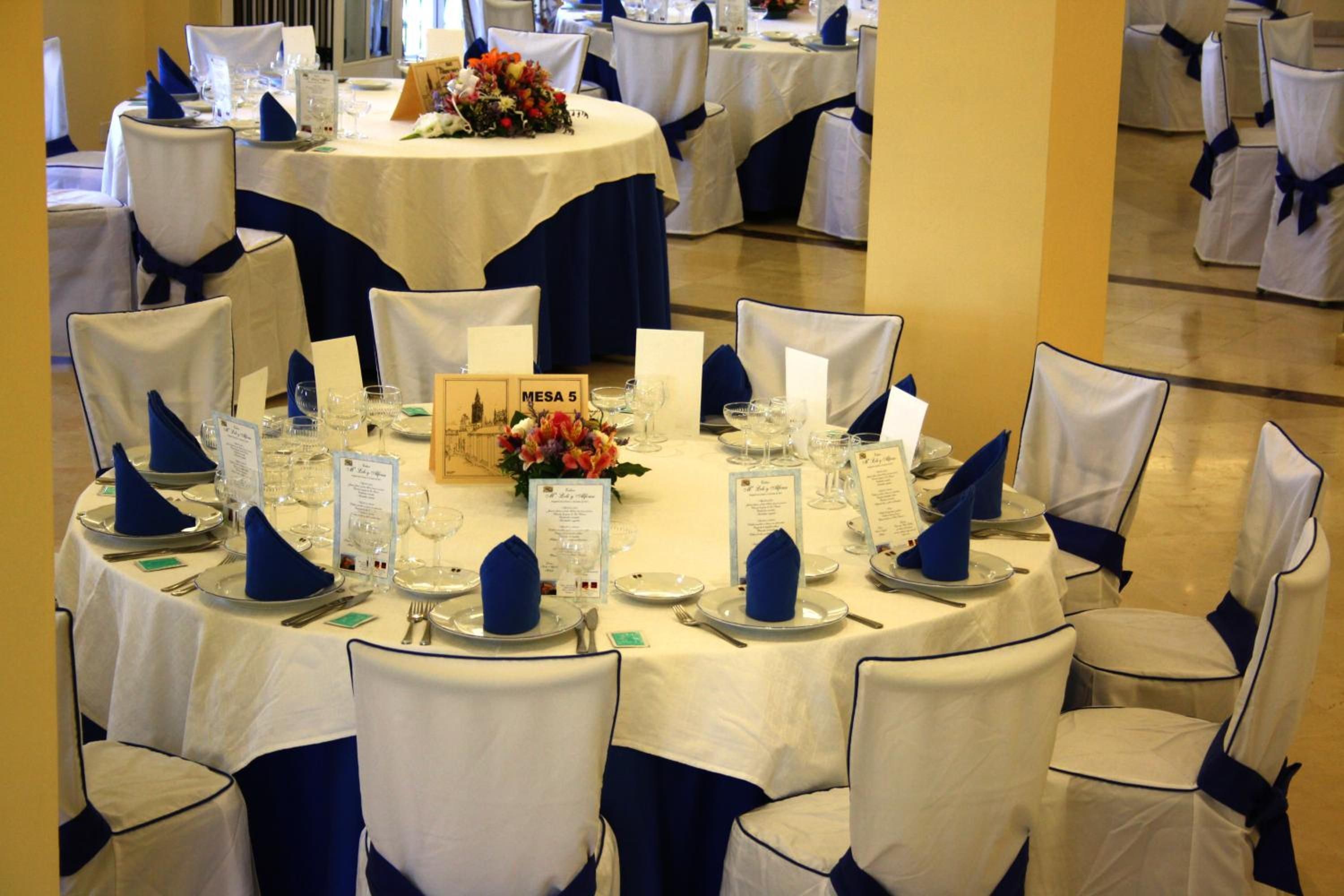 Un salón de banquetes con mesas redondas, manteles blancos y sillas adornadas con cintas azules.