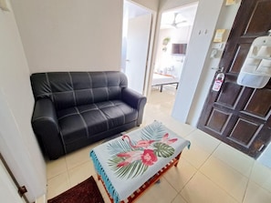 A living room with a black leather sofa, a wooden coffee table with a floral table runner, and a door with a security lock. - Lily holiday  (Patong)