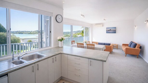 A modern kitchen with a large window, a dining area with a view, and a living space with a sofa and a painting. - Snells Beach Family Retreat — Walk to Shops & Beach (Snells Beach)