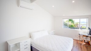 A bedroom with a bed, a desk, a chair, a window with a view, and an air conditioning unit. - Snells Beach Family Retreat — Walk to Shops & Beach (Snells Beach)