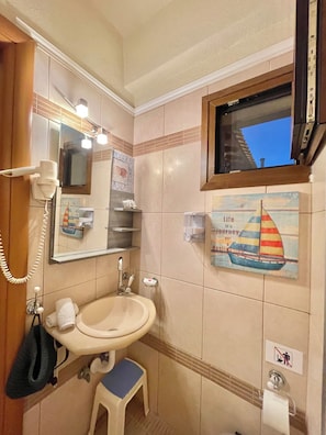 A bathroom with a sink, a mirror, a towel rack, a small stool, a hand dryer, and a window with a view of a building.