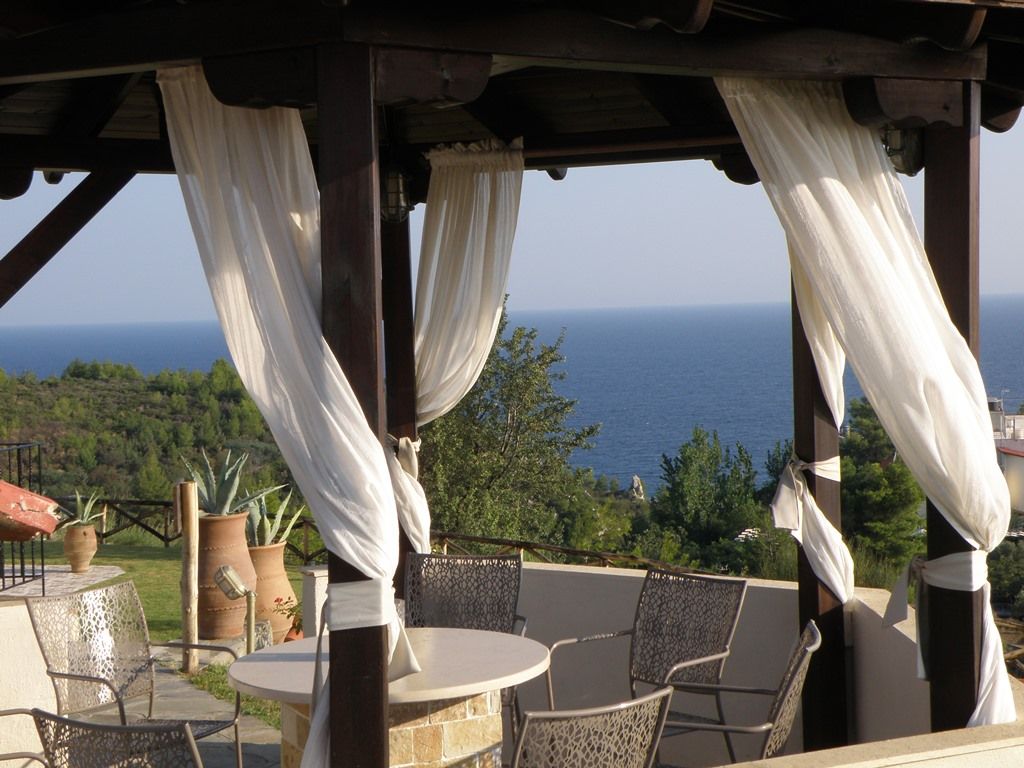 A covered outdoor seating area with a view of the sea, featuring a round table and chairs, and potted plants.