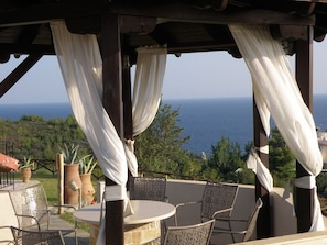 A covered outdoor seating area with a view of the sea, featuring a round table and chairs, and potted plants. - Palamidi Boutique Apartments (Kassandra)