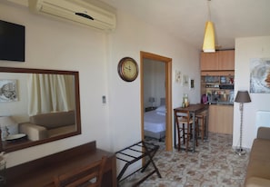 Habitación de hotel con cama, escritorio, silla, espejo, reloj, zona de cocina y mesa de comedor.