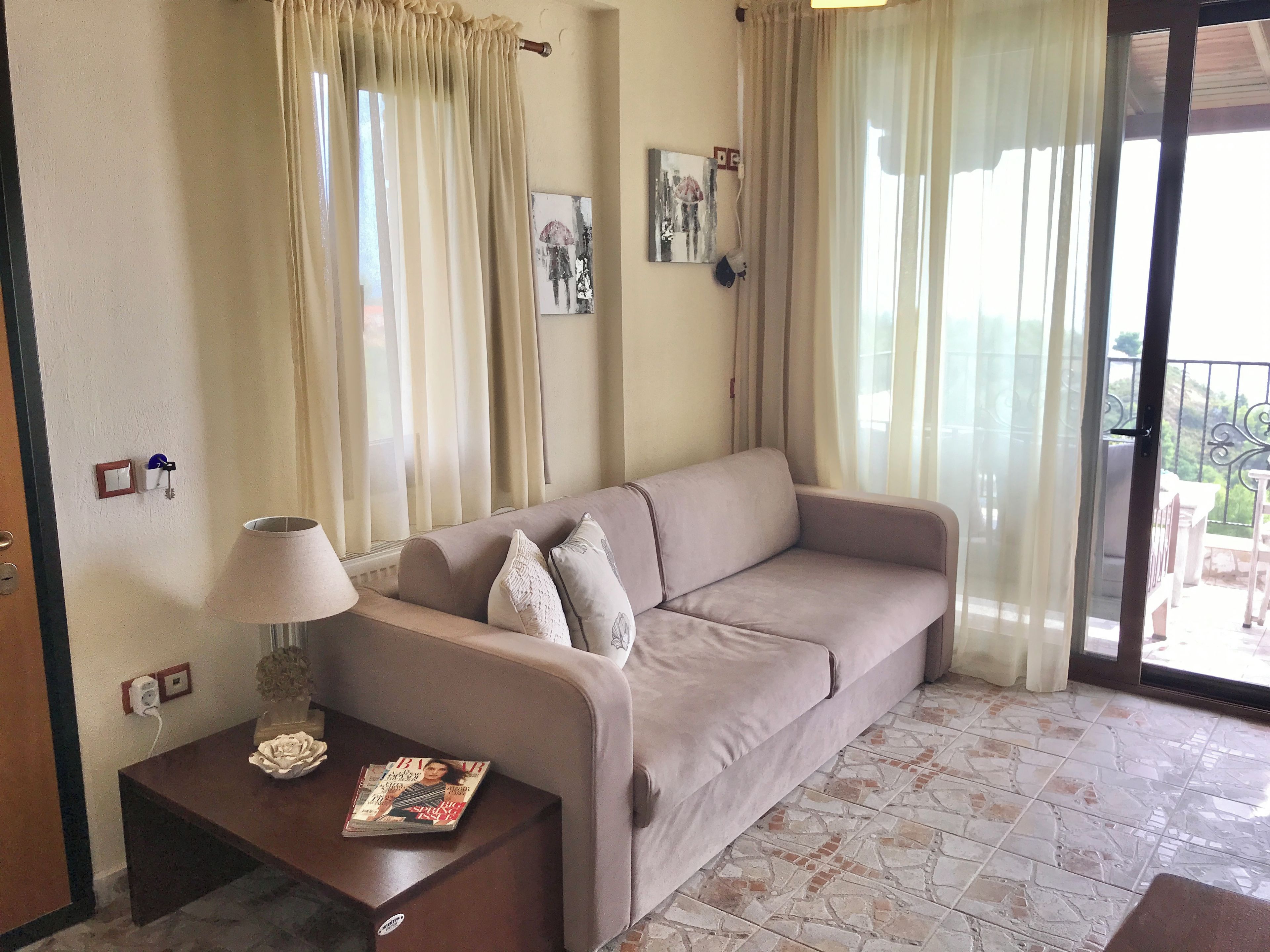 A living room with a beige sofa, a table with a lamp and a magazine, and a view of a balcony through sheer curtains.