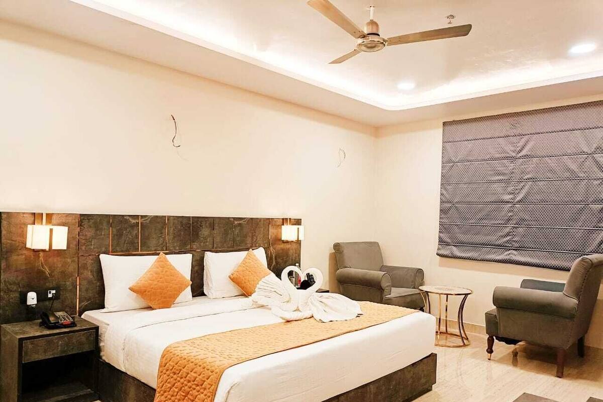 A modern hotel room with a large bed, a ceiling fan, a seating area with a chair and a small table, and a large window with a view.