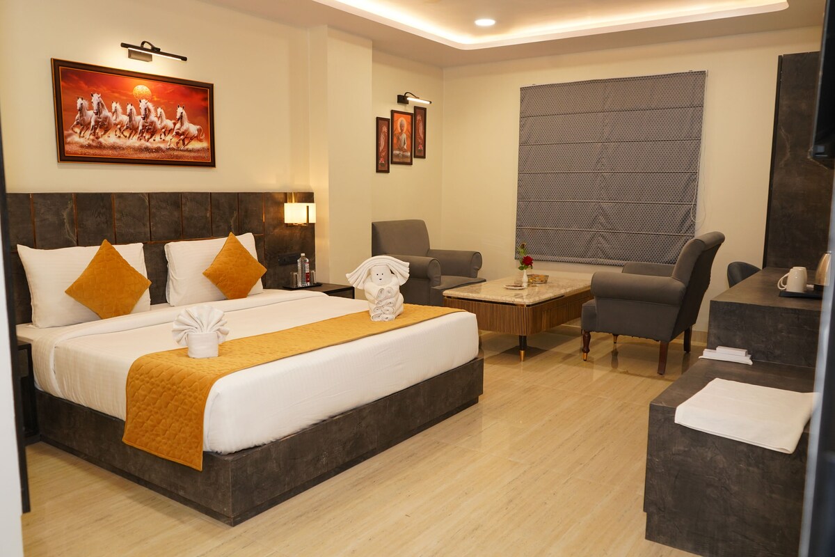 Habitación de hotel con una cama grande, un escritorio, dos sillones, una mesa de centro y un ventanal con persianas.