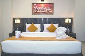 A neatly made bed with a headboard, two bedside lamps, a painting above the bed, and a small nightstand with a bottle and a phone. - Shrigo Hotel Banaras - Varanasi (Varanasi)