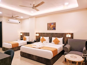 A hotel room with two beds, a sofa, a small table, and a ceiling fan. - Shrigo Hotel Banaras - Varanasi (Varanasi)