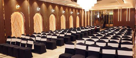 A spacious hall with rows of black chairs and white sashes, a grand chandelier, and a staircase leading to an upper level.