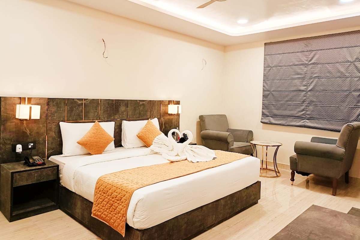 A modern hotel room with a large bed, a ceiling fan, a seating area with a chair and a small table, and a large window with a view.