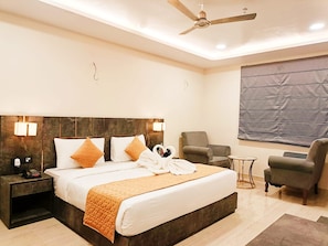 A modern hotel room with a large bed, a ceiling fan, a seating area with a chair and a small table, and a large window with a view. - Shrigo Hotel Banaras - Varanasi (Varanasi)