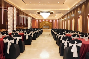 A banquet hall with rows of tables and chairs, a chandelier, and a stage at the front. - Shrigo Hotel Banaras - Varanasi (Varanasi)