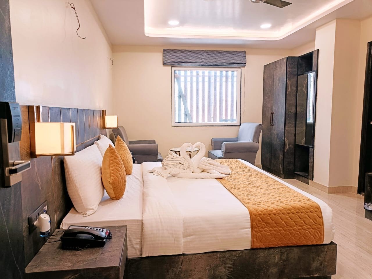 A modern hotel room with a large bed, a bedside table with a phone, a window with blinds, and a TV on a stand.