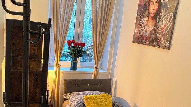 Et soveværelse med seng, et vindue med gardiner, et billede på væggen og et lille bord med en vase blomster.