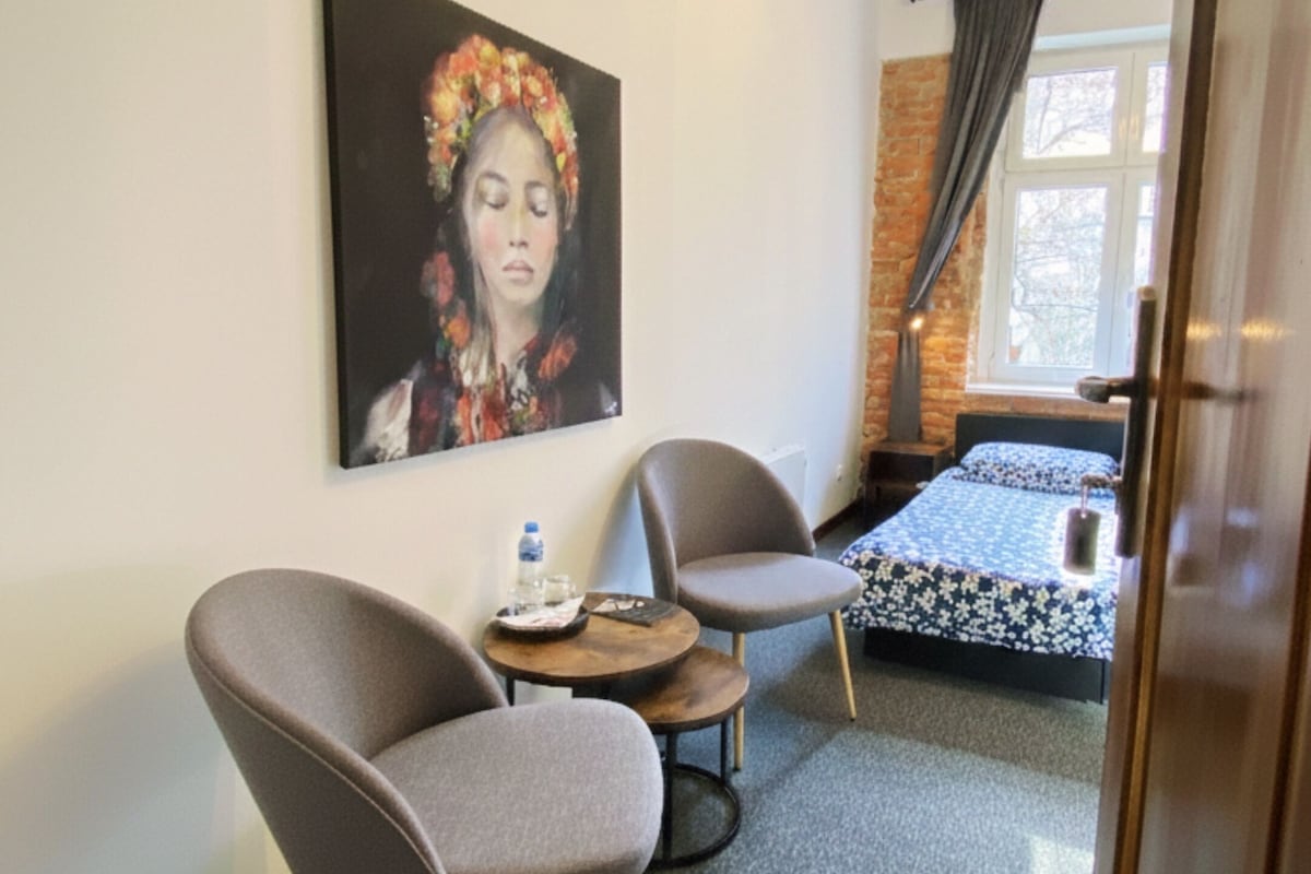 Een kamer met twee grijze fauteuils, een klein rond tafeltje, een bed met een bloemenbedekking, een schilderij aan de muur en een raam met gordijnen.