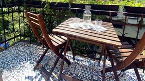 Et træbord og stole på en fliseterrasse med udsigt til grønne områder og en bygning i baggrunden.
