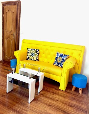 Ein hellgelbes Sofa mit gemusterten Kissen, ein weißer Couchtisch und ein blauer Hocker in einem Raum mit Holzboden und einer Holztür.