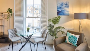 En moderne stue med et glasbord, to stole, en sofa, en gulvlampe og udsigt til bygninger gennem et vindue med skodder.
