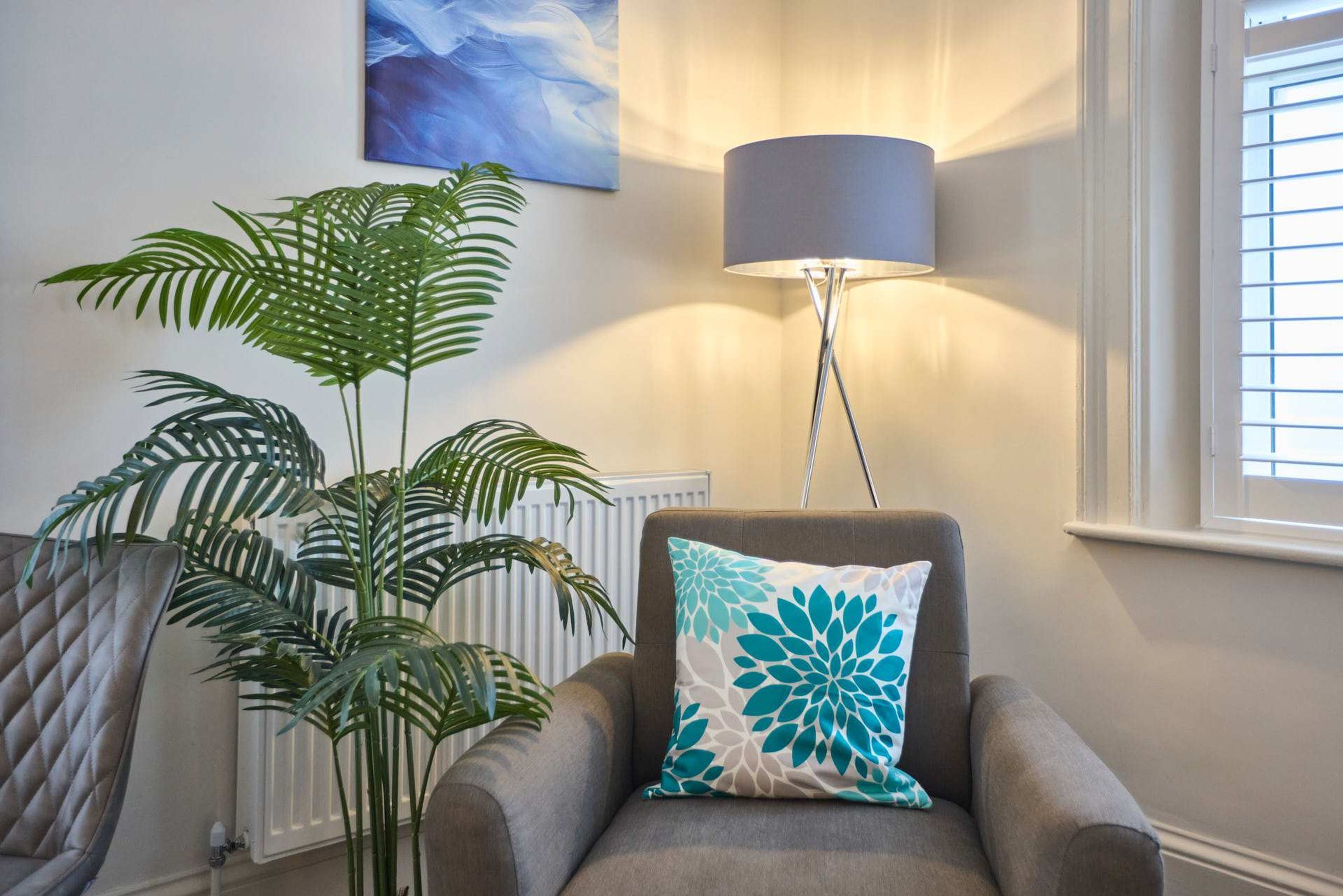 Een zithoek met een grijze fauteuil, een grote potplant, een vloerlamp en een schilderij aan de muur.