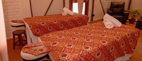 Dos camas individuales con ropa de cama estampada, un taburete de madera y una silla de masaje en una habitación con paredes blancas y una pared de ladrillo decorativa.