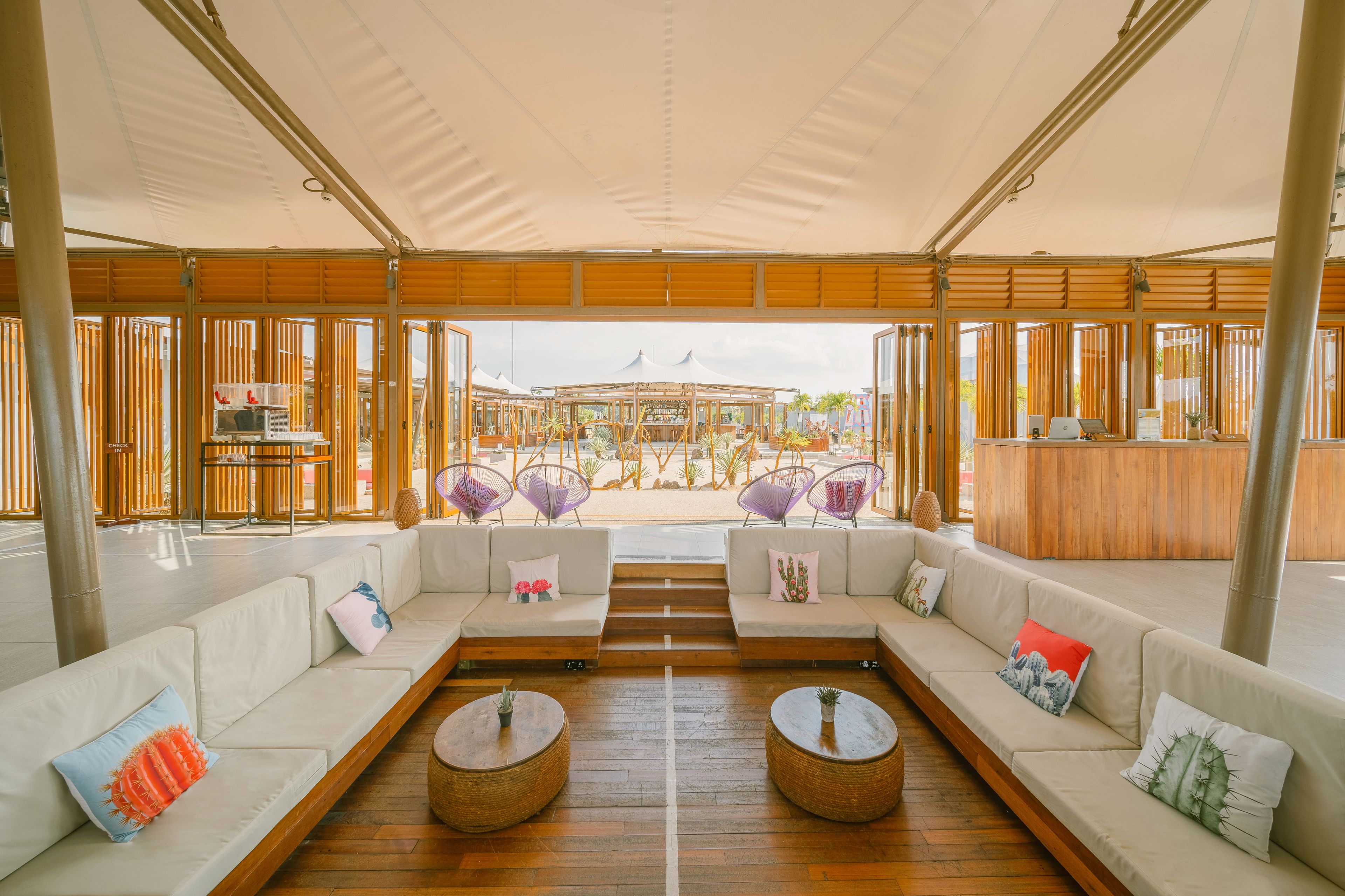 A spacious lounge area with a long white sofa, two round wicker tables, and purple woven chairs.