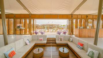 A spacious lounge area with a long white sofa, two round wicker tables, and purple woven chairs.
