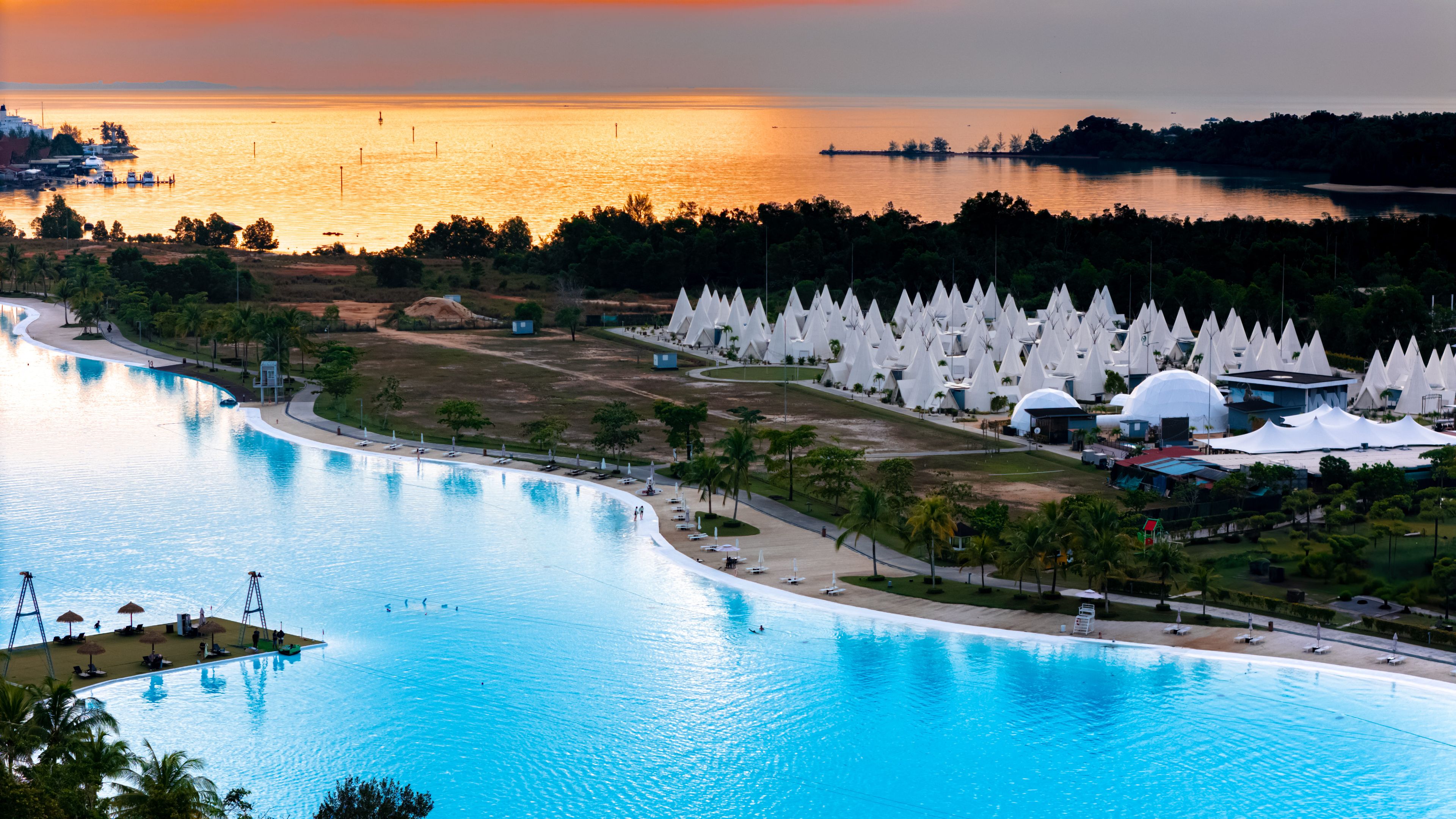 A large swimming pool with a curved edge, surrounded by a landscaped area with palm trees and a beach.