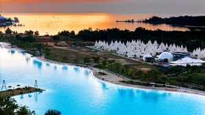 A large swimming pool with a curved edge, surrounded by a landscaped area with palm trees and a beach. - The Anmon Resort Bintan (Bintan)