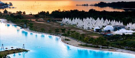 A large swimming pool with a curved edge, surrounded by a landscaped area with palm trees and a beach.
