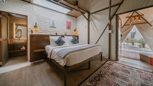 A tented accommodation room with a bed, a wooden headboard, a white brick wall, a skylight, a small table with a lamp, and a view of the outside. - The Anmon Resort Bintan (Bintan)