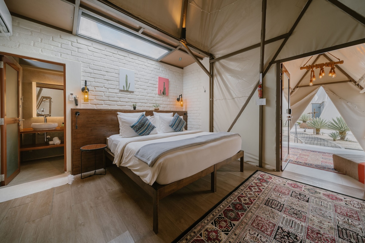 A tented accommodation room with a bed, a wooden headboard, a white brick wall, a skylight, a small table with a lamp, and a view of the outside.