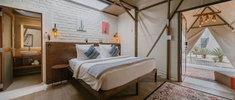 Une chambre aménagée sous une tente, comprenant un lit, une tête de lit en bois, un mur en briques blanches, une lucarne, une petite table avec une lampe et une vue sur l’extérieur.
