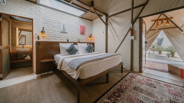 A tented accommodation room with a bed, a wooden headboard, a white brick wall, a skylight, a small table with a lamp, and a view of the outside. - The Anmon Resort Bintan (Bintan)