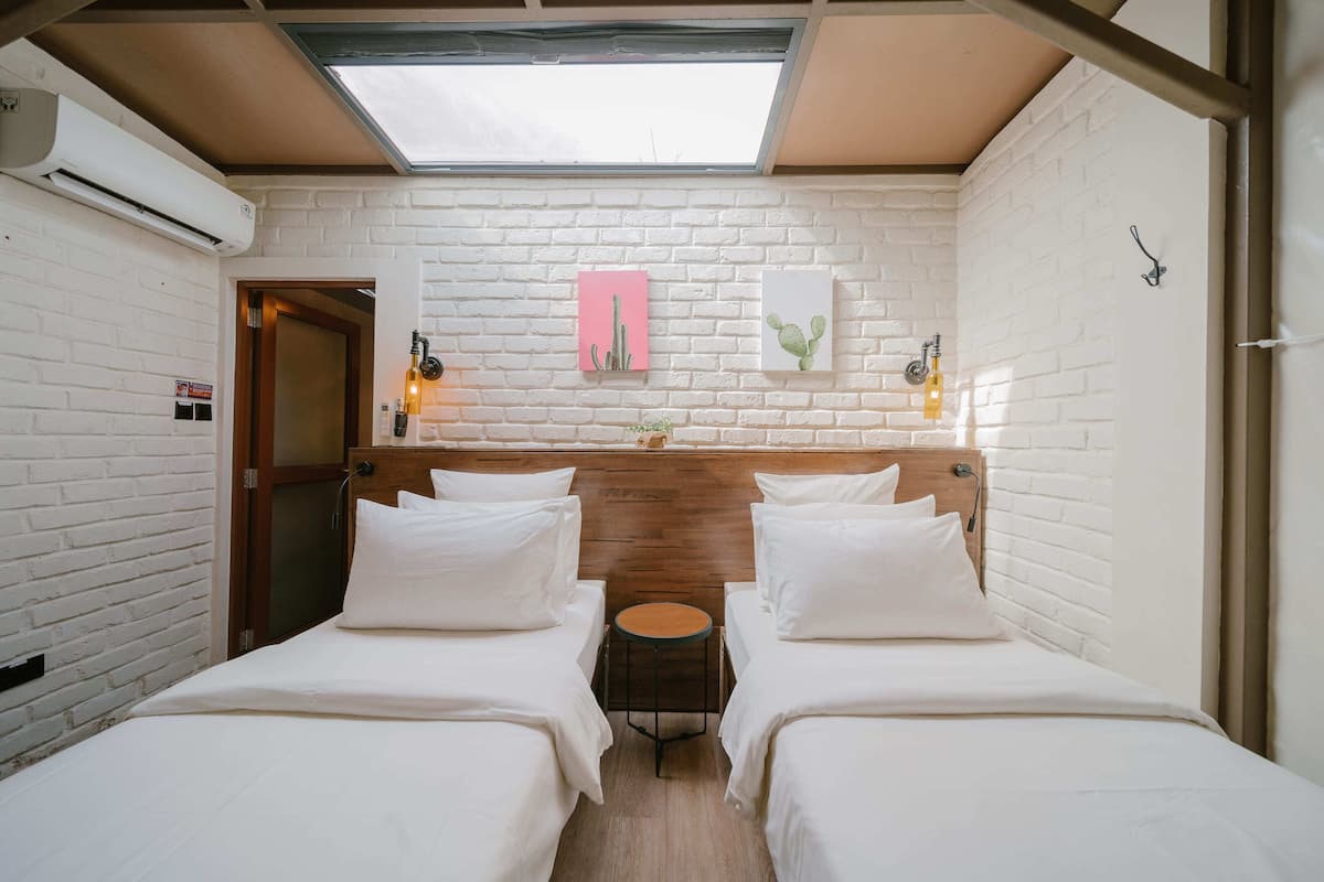 Una habitación con dos camas, un tragaluz, paredes de ladrillo blanco y un cabecero de madera.