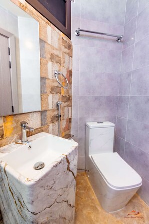 A bathroom with a marble sink, a toilet, and a shower area. - Desert Rose Village (Abu Dhabi)