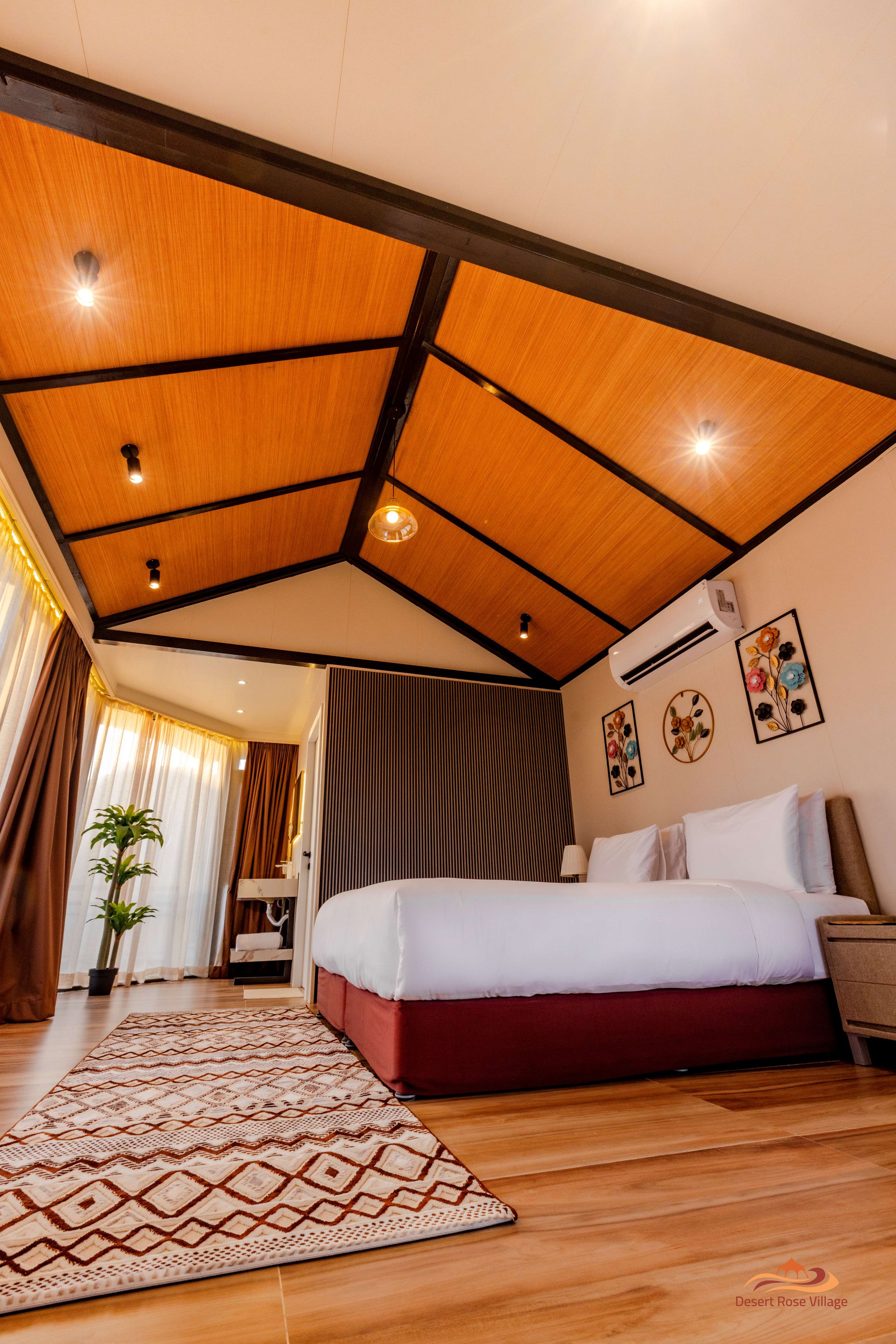 A hotel room with a wooden ceiling, a bed with white and red bedding, a patterned rug, and a potted plant.