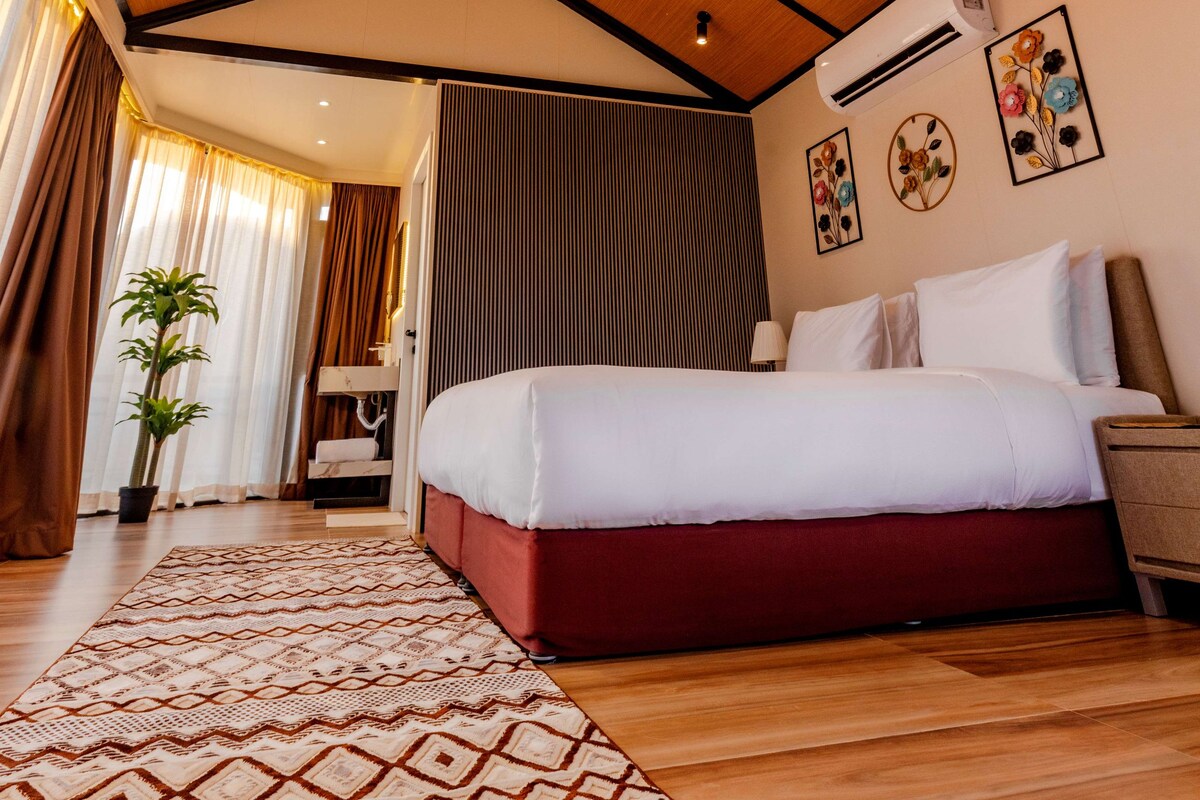 Habitación de hotel con techo de madera, cama con ropa de cama blanca y roja, alfombra estampada y planta en maceta.