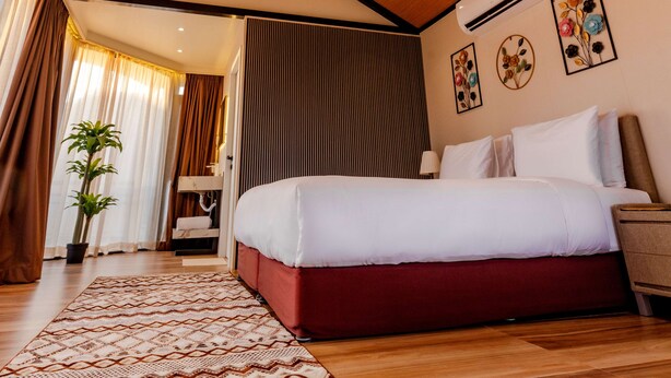 A hotel room with a wooden ceiling, a bed with white and red bedding, a patterned rug, and a potted plant.
