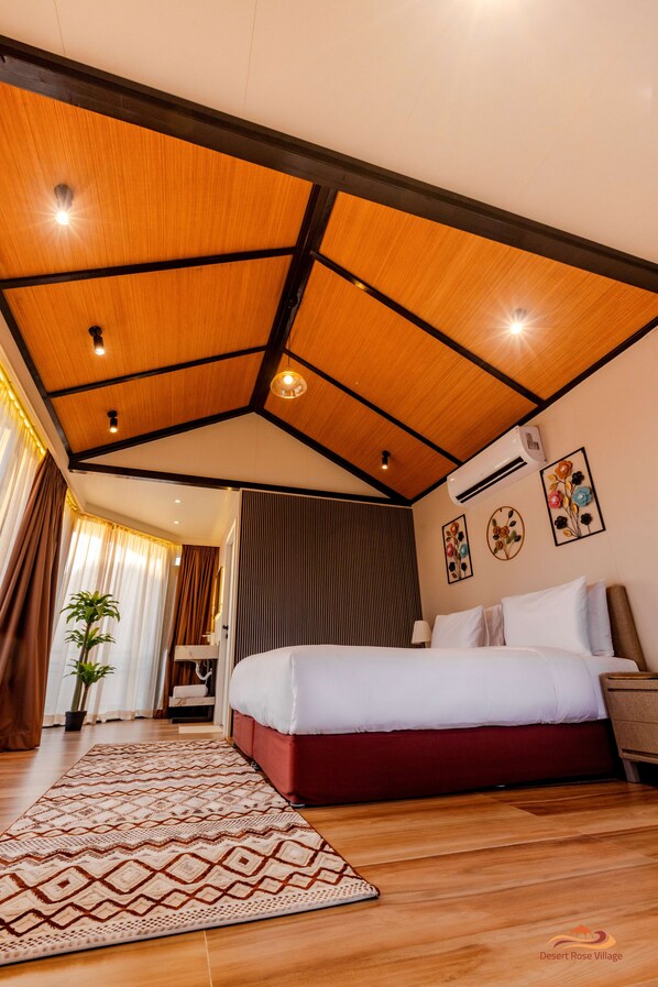 Habitación de hotel con techo de madera, cama con ropa de cama blanca y roja, alfombra estampada y planta en maceta.