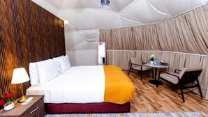 A room with a bed, a bedside table with a lamp, a chair, and a small table with a plant.
