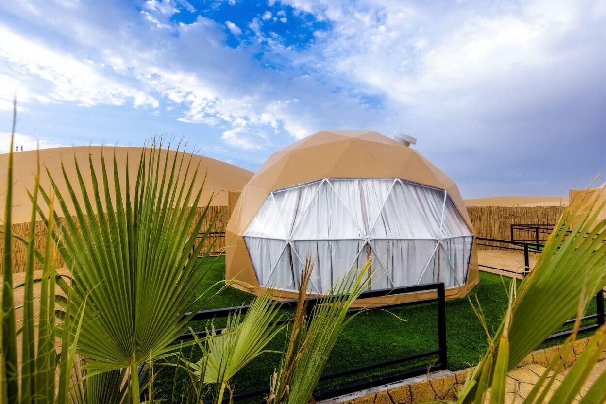 Une structure en forme de dôme, recouverte d’un rideau blanc, entourée de palmiers et surplombée par un ciel dégagé.