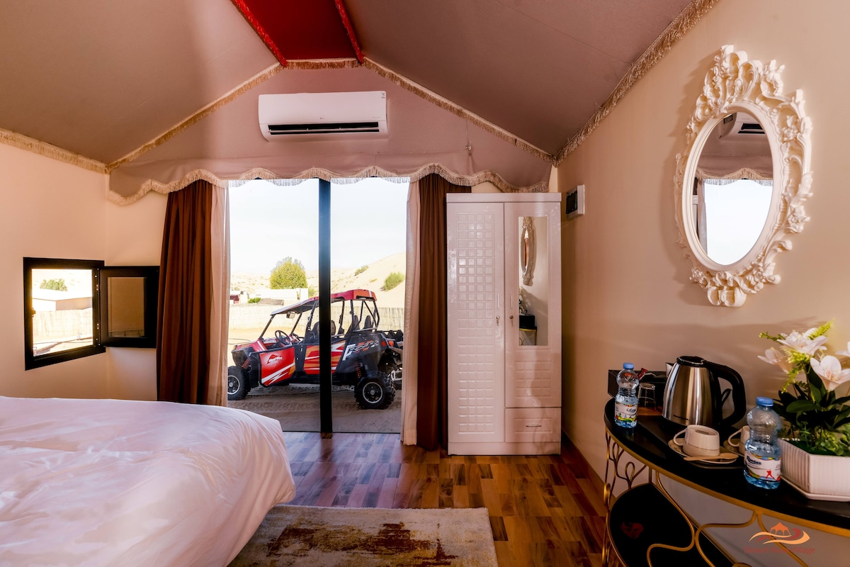 Un dormitorio con cama, tocador con espejo, escritorio con hervidor y vista a un vehículo rojo en el exterior.