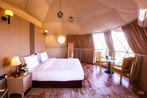 Una habitación tipo tienda de campaña con una cama, mesita de noche, lámpara y una pequeña mesa con sillas.