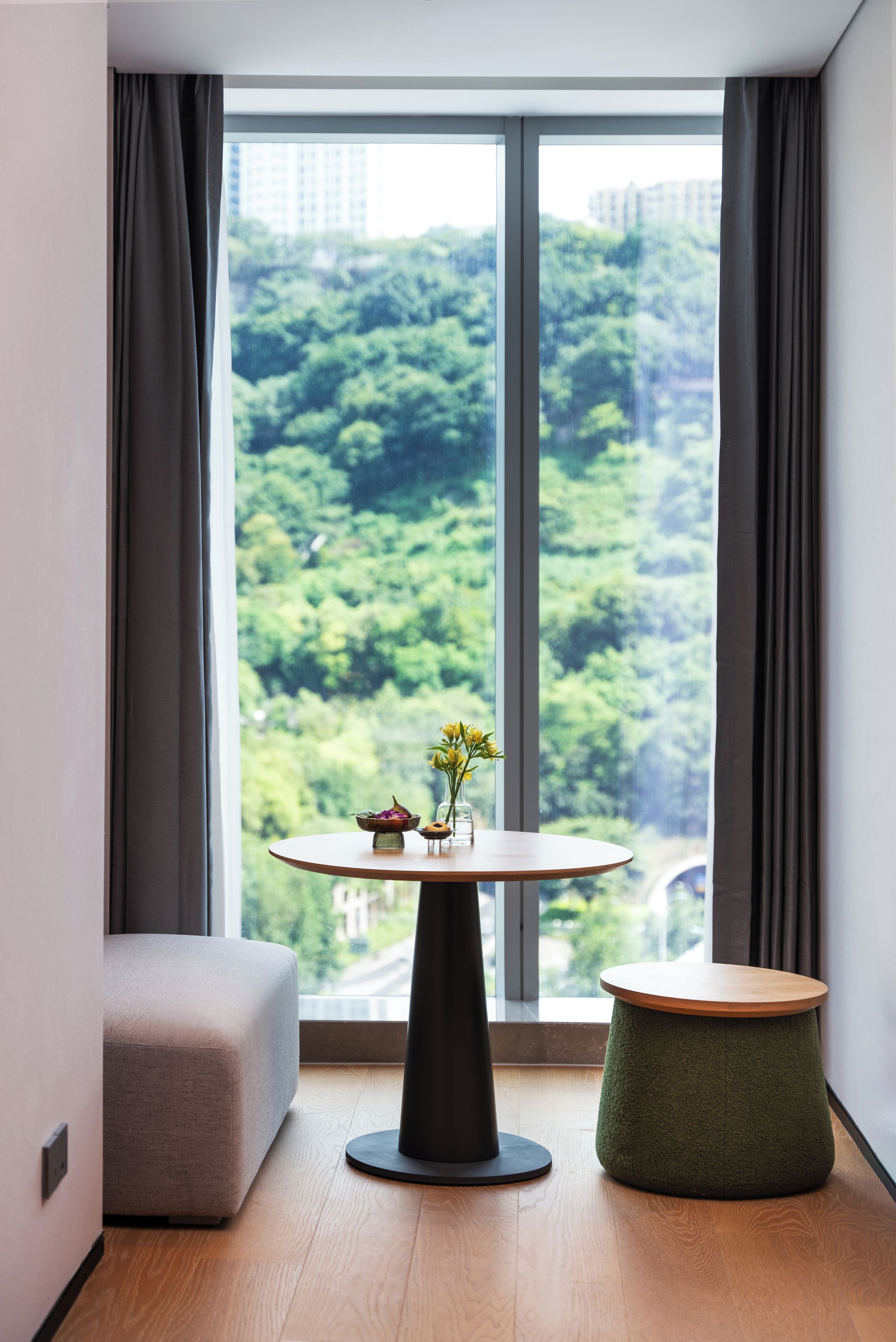 A modern room with a large window, a round table with a vase of flowers, a grey sofa, and a green stool.
